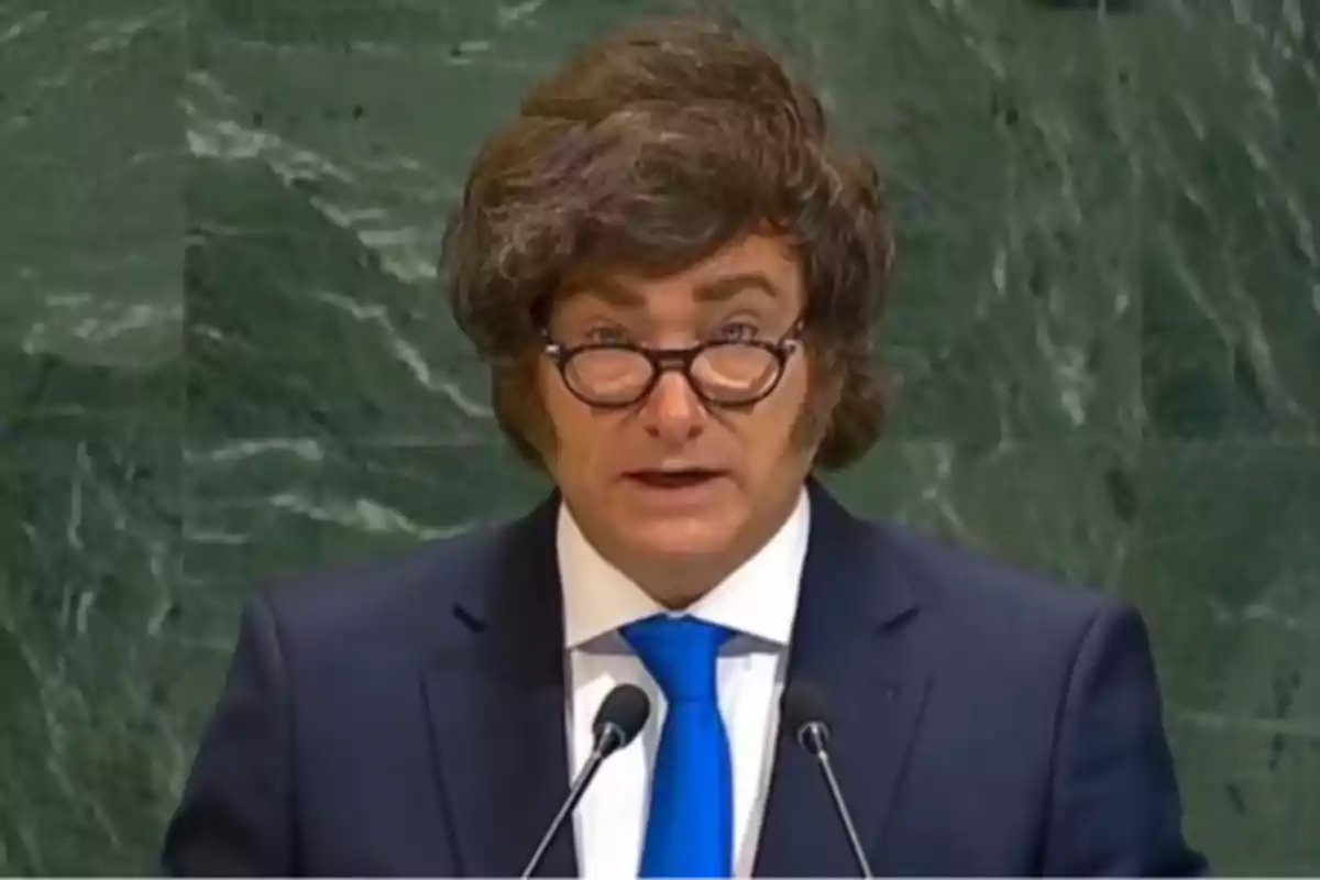Hombre con gafas y cabello voluminoso usando traje azul y corbata azul hablando frente a dos micrófonos con fondo de mármol verde