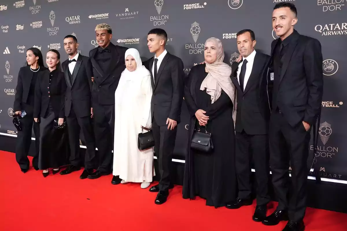 Un grupo de personas elegantemente vestidas posa sobre una alfombra roja frente a un fondo con logotipos de un evento de gala