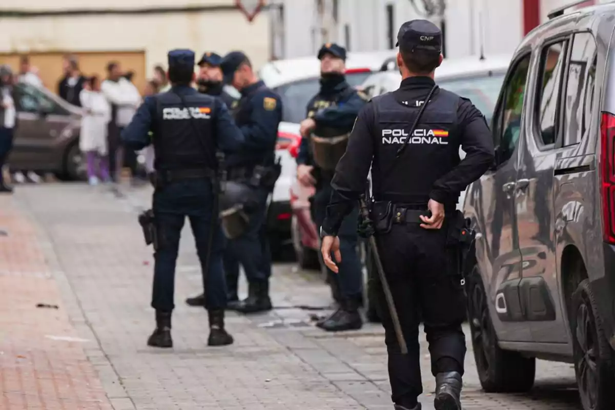Varios agentes de la Policía Nacional patrullan una calle mientras un grupo de personas observa a lo lejos