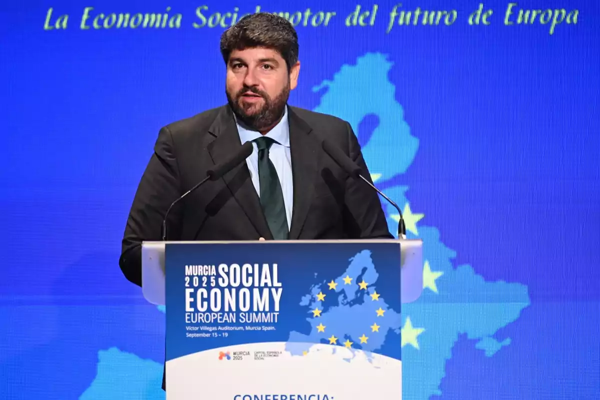 Hombre con barba y traje oscuro hablando en un podio durante una cumbre europea de economía social en Murcia con un fondo azul y un mapa de Europa con estrellas amarillas.