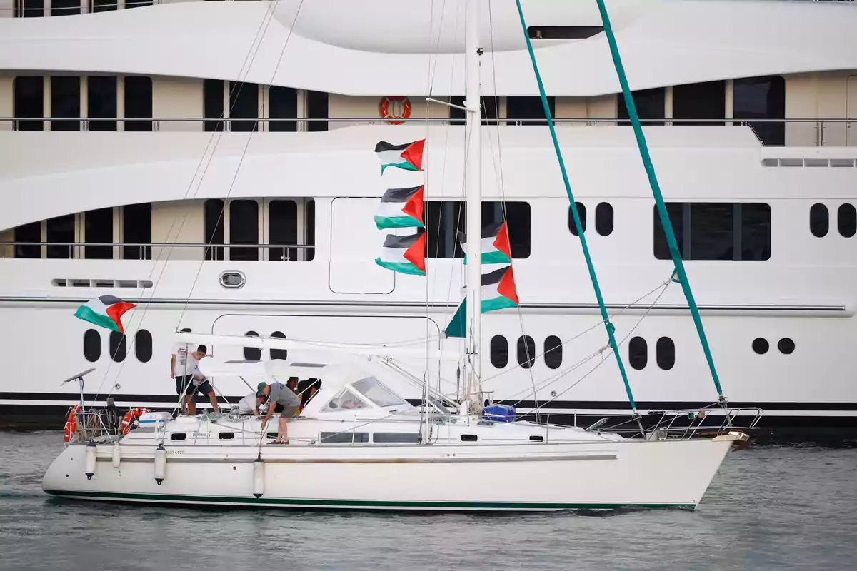 Un velero blanco con varias banderas palestinas navega frente a un gran yate de lujo blanco mientras varias personas están a bordo del velero Un velero blanco con varias banderas palestinas navega frente a un gran yate de lujo blanco mientras varias personas están a bordo del velero