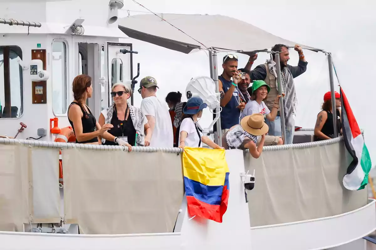 Personas en la cubierta de un barco con una bandera de Colombia y una bandera de Palestina colgadas en la barandilla, algunas personas conversan y otras observan el paisaje Personas en la cubierta de un barco con una bandera de Colombia y una bandera de Palestina colgadas en la barandilla, algunas personas conversan y otras observan el paisaje