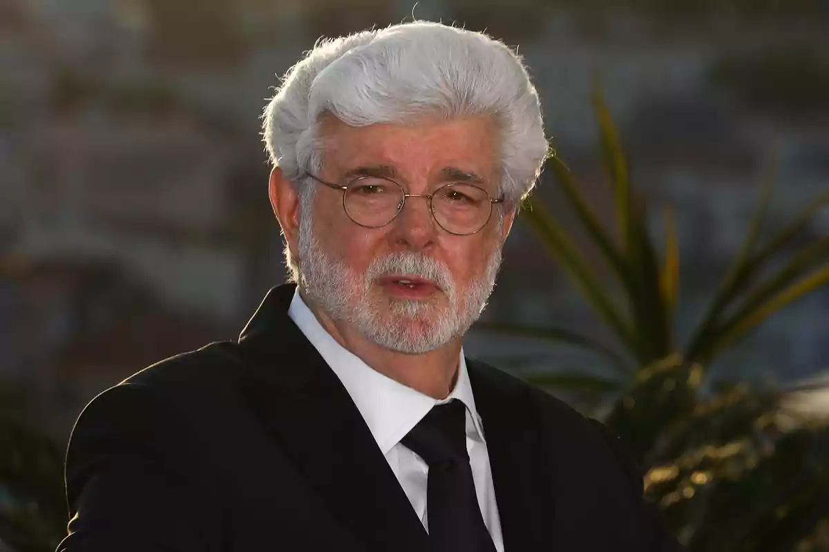 George Lucas de cabello y barba blanca con gafas y traje oscuro posando al aire libre con plantas y fondo desenfocado