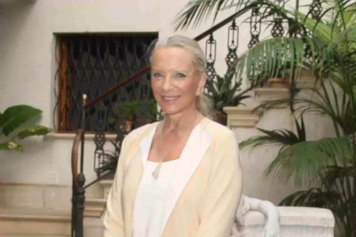 La duquesa de Kent sonriendo, vestida con ropa clara, posando en un ambiente interior con plantas y una escalera al fondo