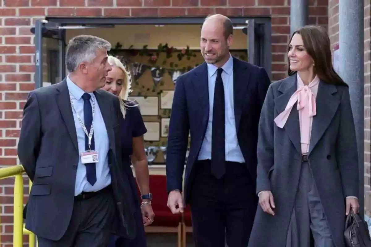 Kate Middleton y el príncipe Guillermo elegantemente vestidos caminan y conversan sonrientes frente a un edificio de ladrillo rojo. Kate Middleton y el príncipe Guillermo elegantemente vestidos caminan y conversan sonrientes frente a un edificio de ladrillo rojo.