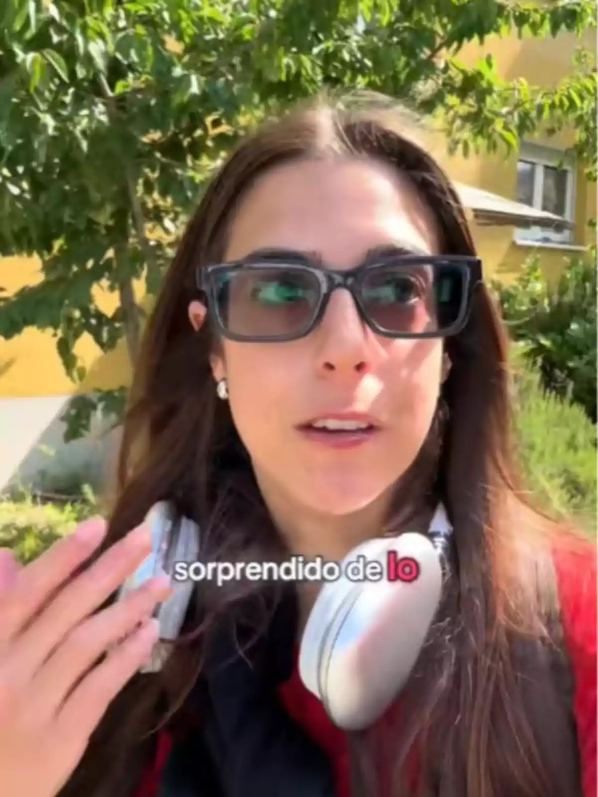 Mujer joven con gafas oscuras y auriculares grandes alrededor del cuello, al aire libre con árboles y un edificio amarillo de fondo, texto en pantalla que dice sorprendido de lo.