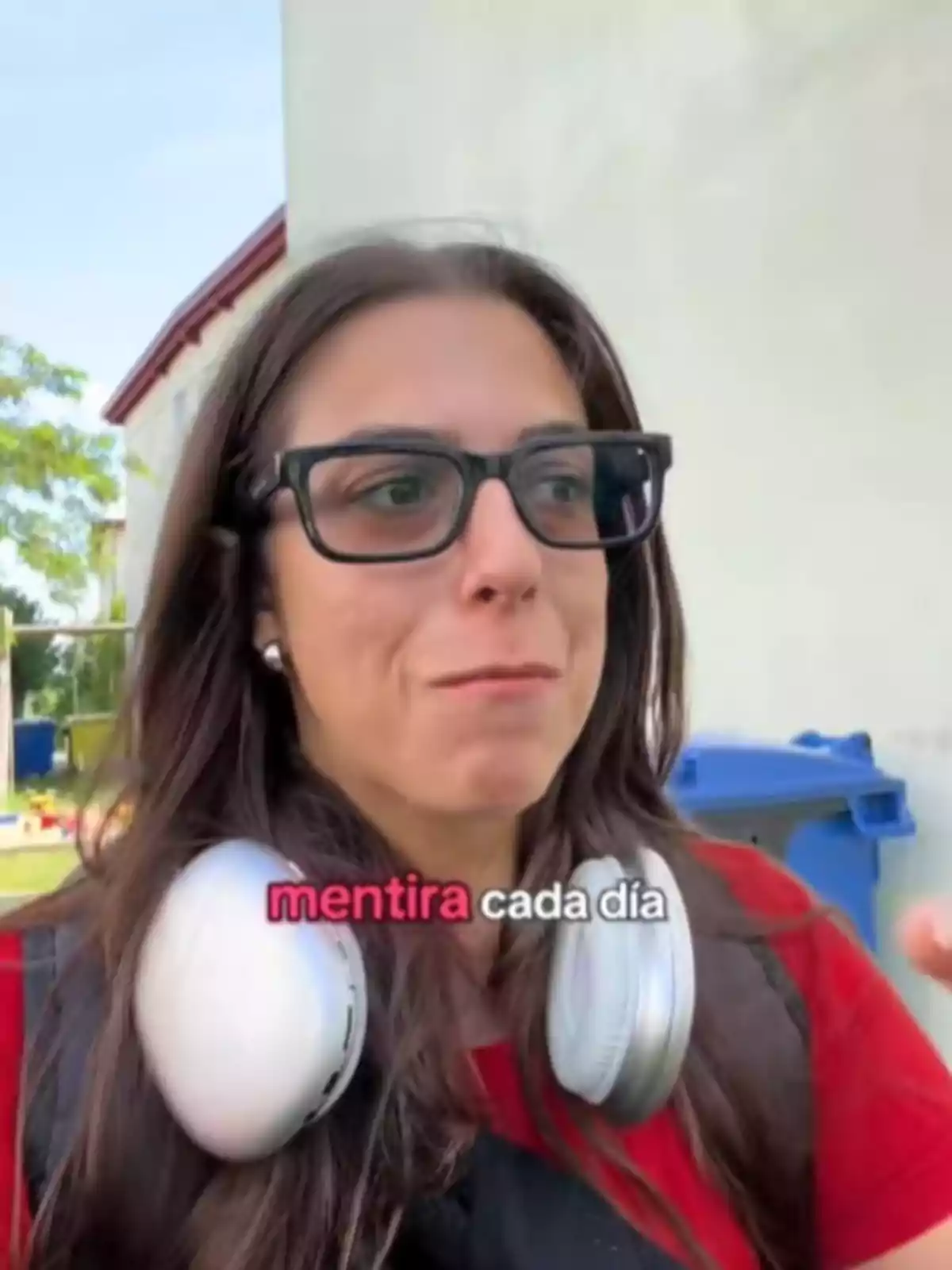 Mujer de cabello largo y gafas negras con audífonos grandes alrededor del cuello, lleva puesta una camiseta roja y aparece el texto mentira cada día sobre la imagen.