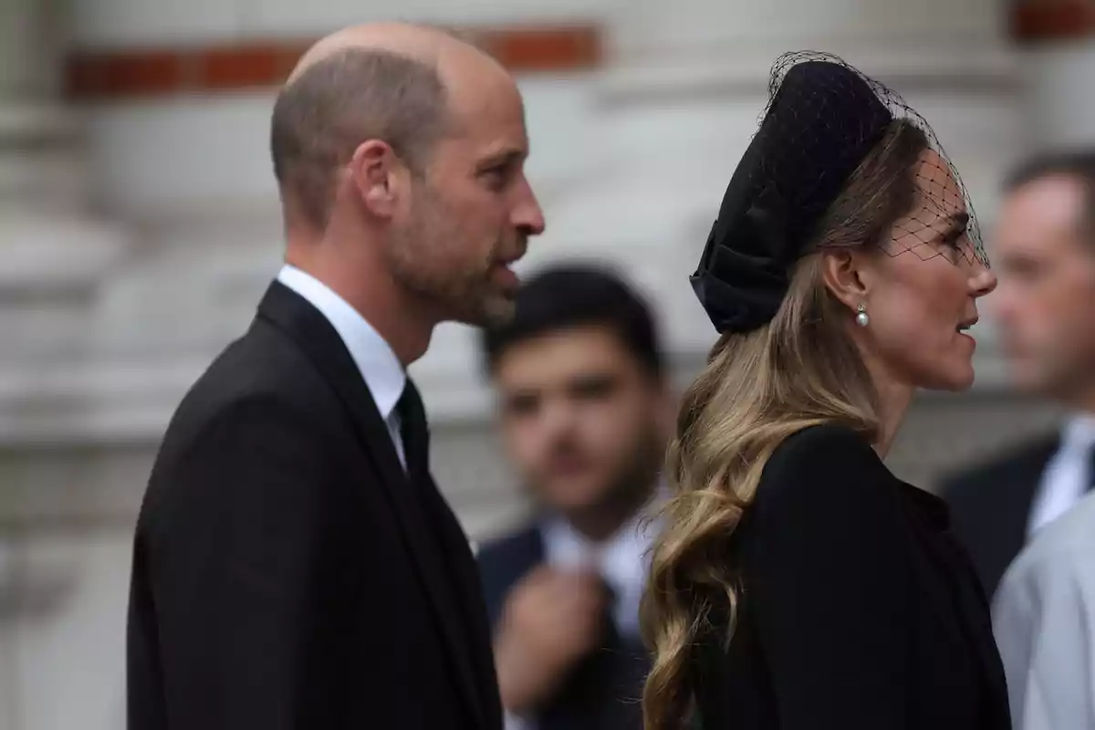Kate Middleton y el príncipe Guillermo vestidos de negro caminan uno al lado de la otra en un evento formal; Kate lleva un tocado negro con velo y pendientes de perla.