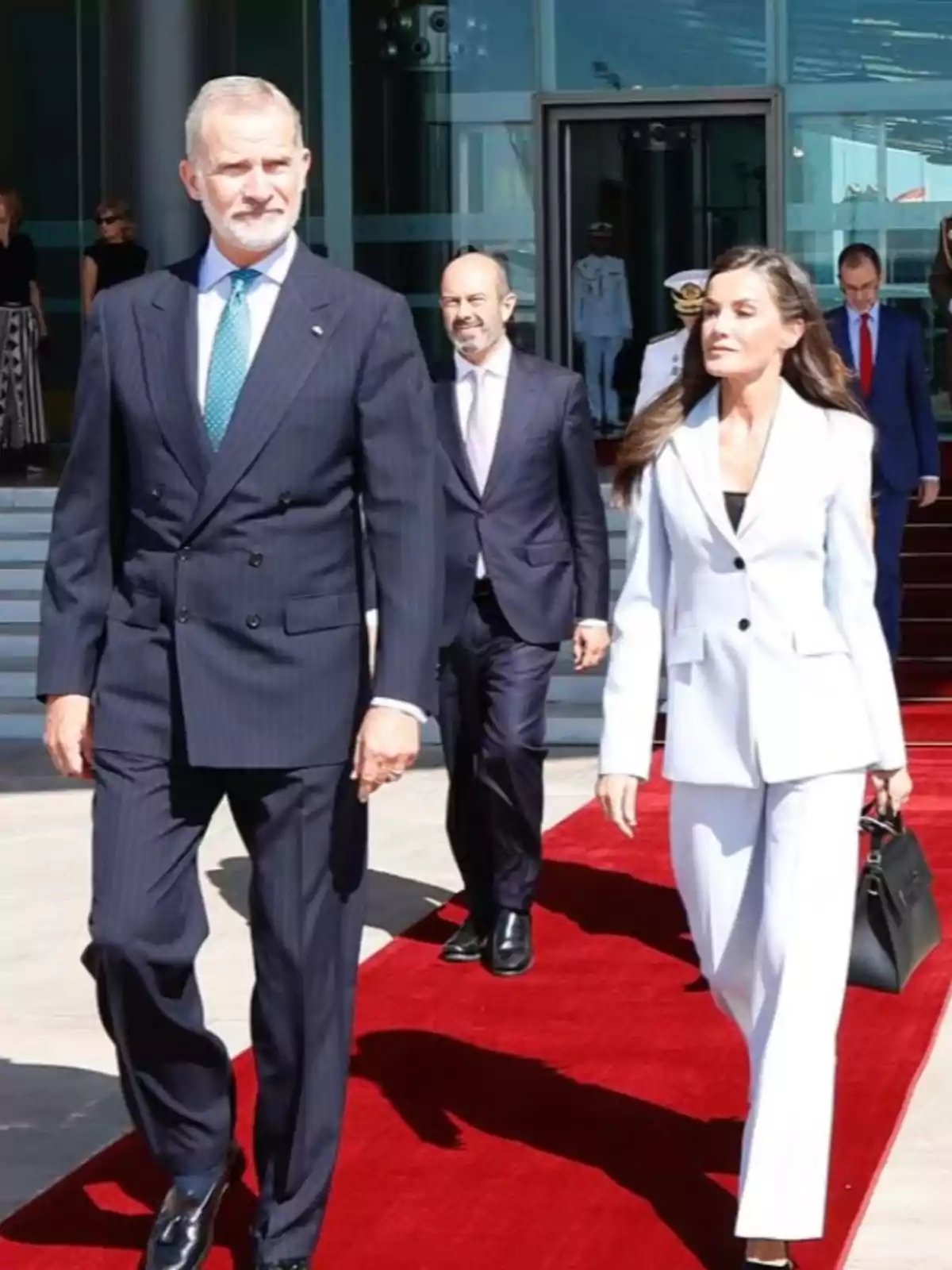 El rey Felipe y la reina Letizia elegantemente vestidos caminan sobre una alfombra roja frente a un edificio moderno, acompañados por otras personas al fondo.