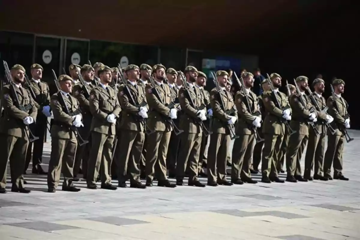 Un grupo de soldados uniformados con guantes blancos y boinas verdes está formado en fila sosteniendo rifles durante una ceremonia al aire libre.