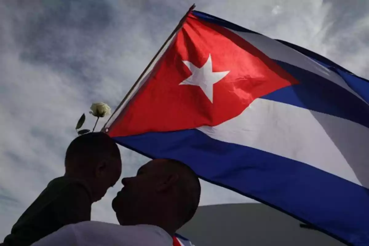 Un hombre carga a un niño que sostiene una rosa blanca mientras una bandera de Cuba ondea al fondo bajo un cielo nublado. Un hombre carga a un niño que sostiene una rosa blanca mientras una bandera de Cuba ondea al fondo bajo un cielo nublado.