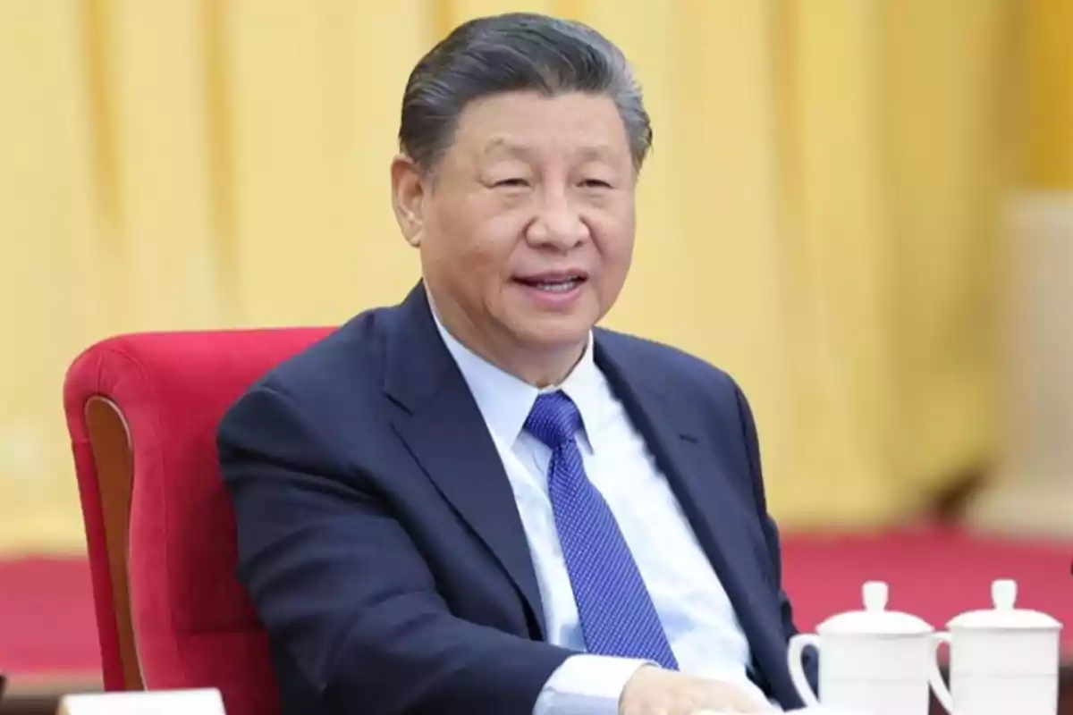 Hombre de cabello canoso y traje azul sentado en una silla roja junto a una mesa con tazas blancas y fondo amarillo Hombre de cabello canoso y traje azul sentado en una silla roja junto a una mesa con tazas blancas y fondo amarillo