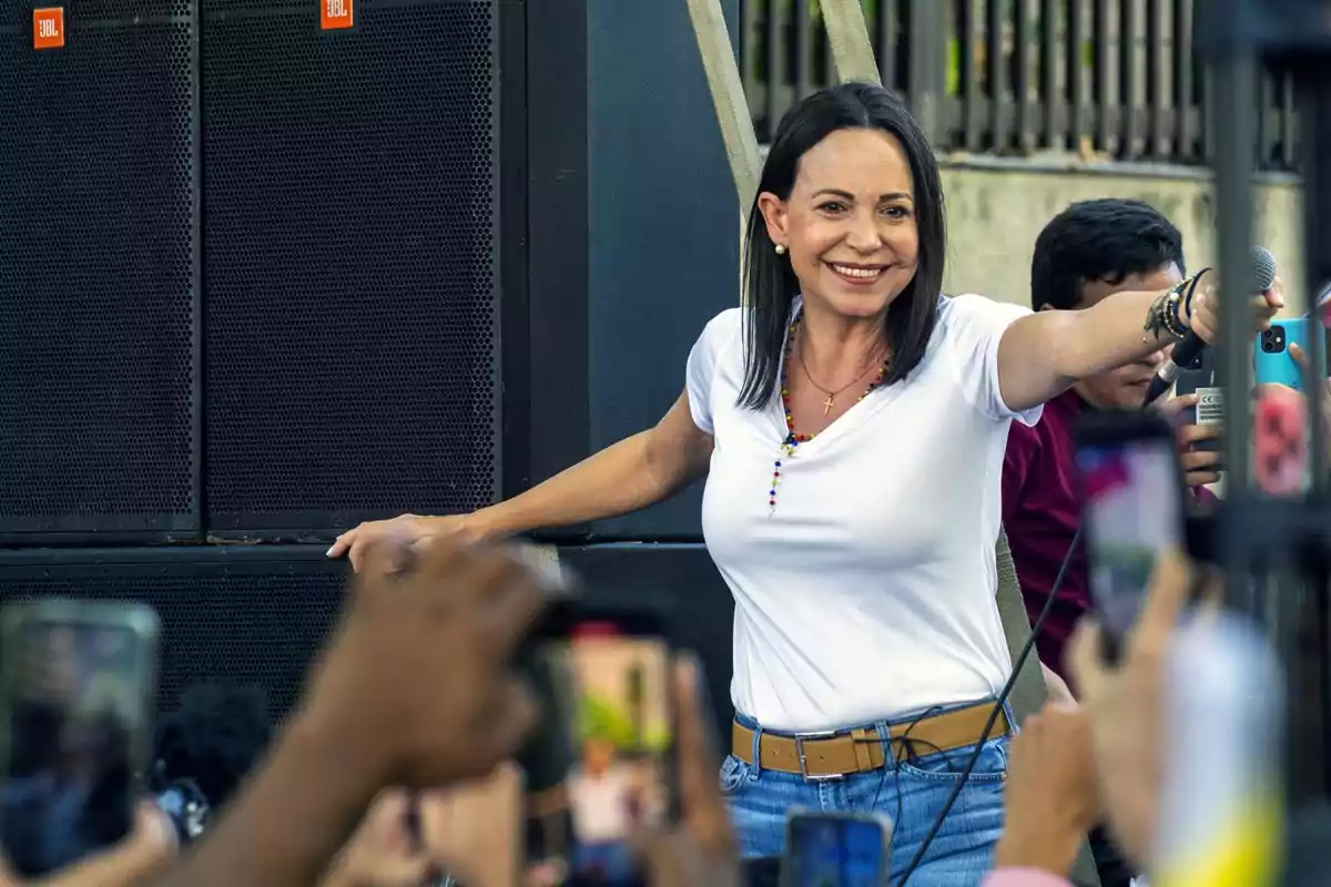 Mujer sonriente con camiseta blanca y collar colorido sostiene un micrófono mientras varias personas la fotografían con sus teléfonos en un evento al aire libre Mujer sonriente con camiseta blanca y collar colorido sostiene un micrófono mientras varias personas la fotografían con sus teléfonos en un evento al aire libre