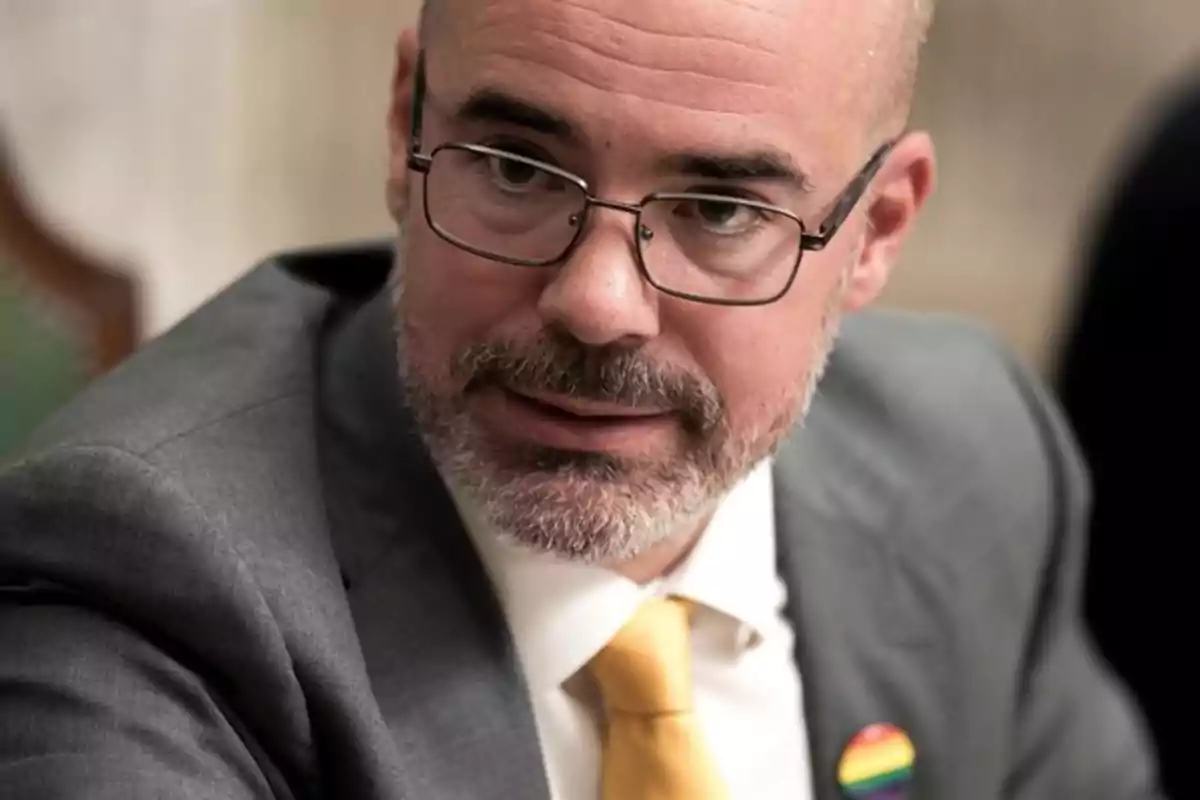 Hombre con gafas y barba vestido con traje gris y corbata amarilla lleva un pin con la bandera arcoíris en la solapa Hombre con gafas y barba vestido con traje gris y corbata amarilla lleva un pin con la bandera arcoíris en la solapa