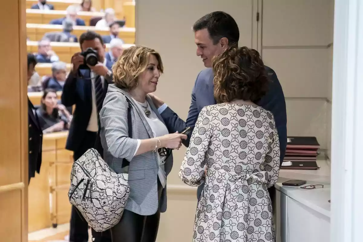 Tres personas conversan en un pasillo mientras al fondo se observa a un fotógrafo y varias personas sentadas en un auditorio Tres personas conversan en un pasillo mientras al fondo se observa a un fotógrafo y varias personas sentadas en un auditorio
