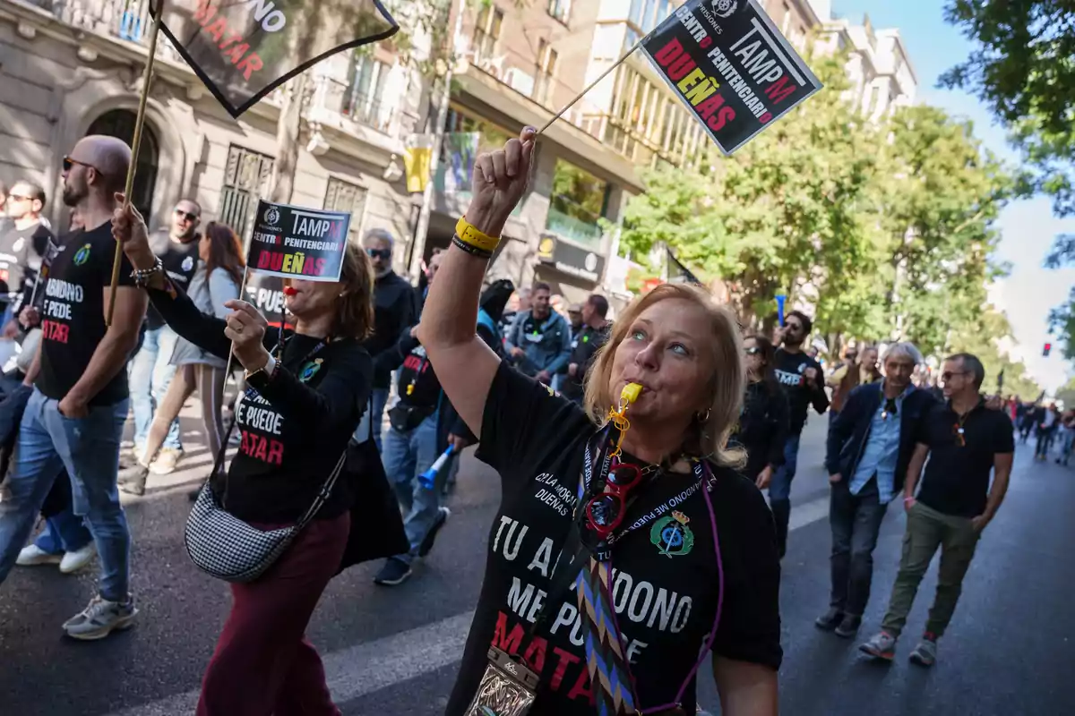 Mujeres y hombres marchan por una calle con camisetas negras y pancartas mientras una mujer al frente sopla un silbato amarillo