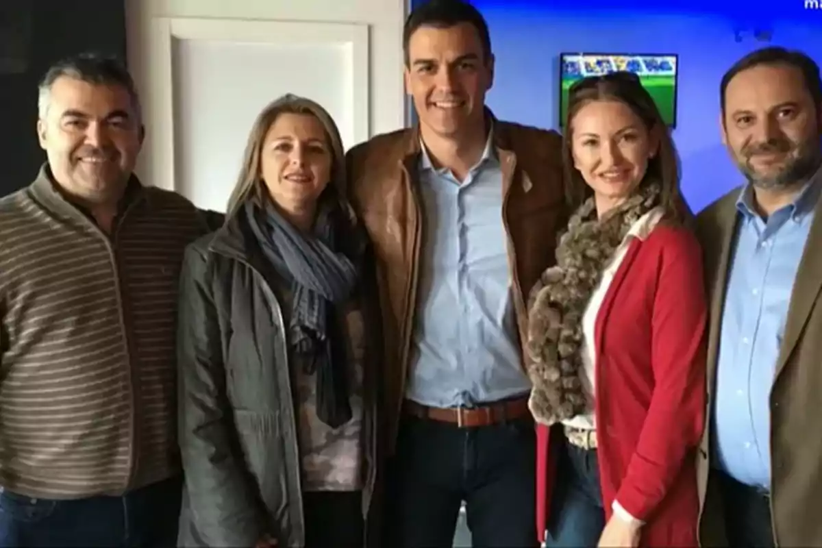 Cinco personas posan juntas y sonríen en un ambiente interior con una pantalla de televisión al fondo. Cinco personas posan juntas y sonríen en un ambiente interior con una pantalla de televisión al fondo.