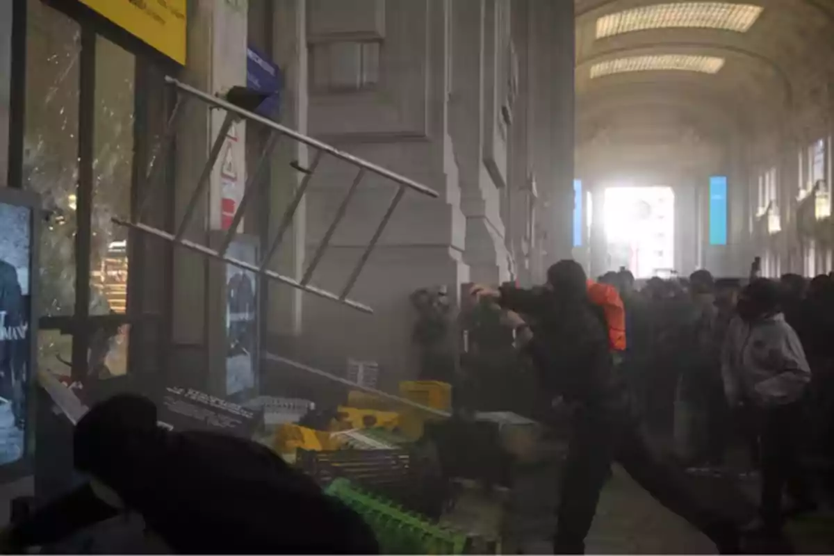 Personas lanzan una escalera contra una puerta de vidrio mientras una multitud observa en el interior de un edificio grande y luminoso Personas lanzan una escalera contra una puerta de vidrio mientras una multitud observa en el interior de un edificio grande y luminoso