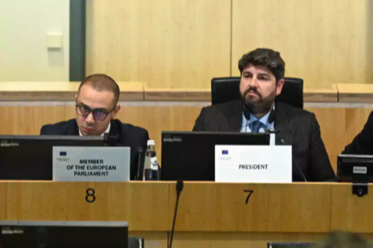 Dos hombres con traje están sentados en una sala de conferencias detrás de escritorios numerados con letreros que indican sus cargos uno es miembro del Parlamento Europeo y el otro es presidente Dos hombres con traje están sentados en una sala de conferencias detrás de escritorios numerados con letreros que indican sus cargos uno es miembro del Parlamento Europeo y el otro es presidente
