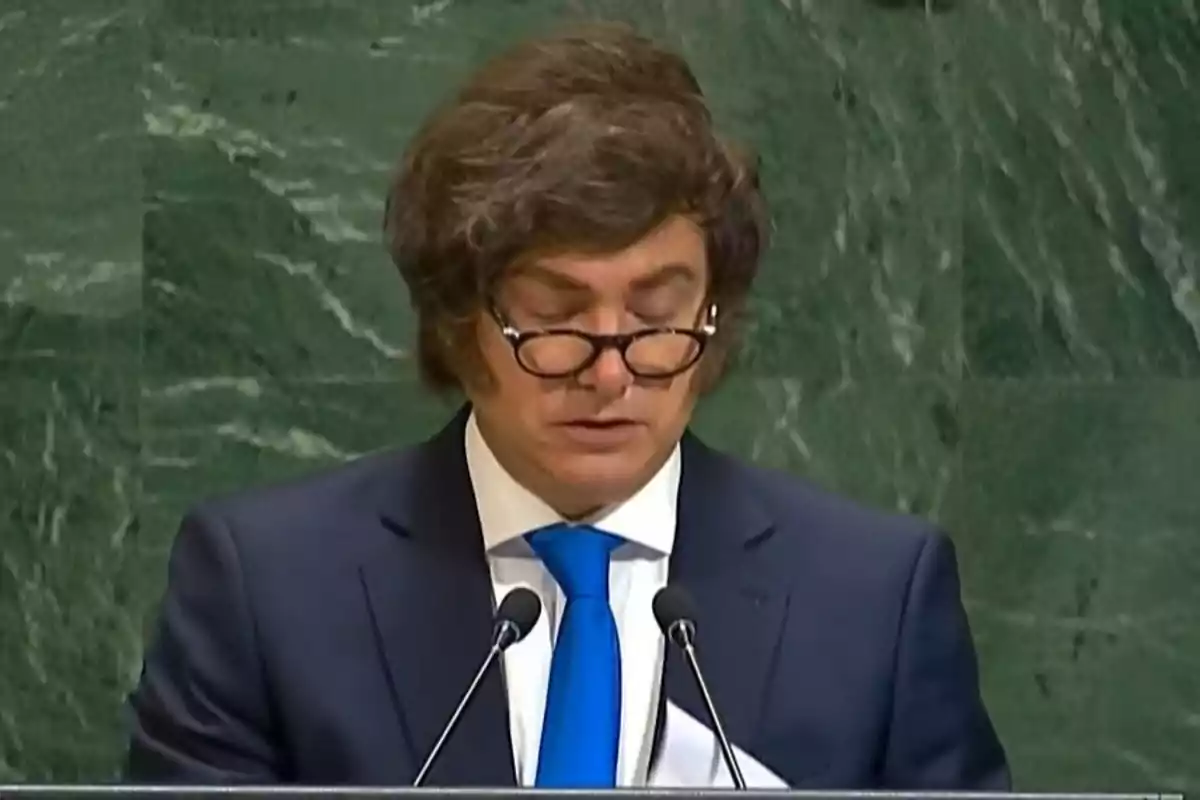 Hombre de cabello castaño y gafas hablando en un podio con fondo de mármol verde