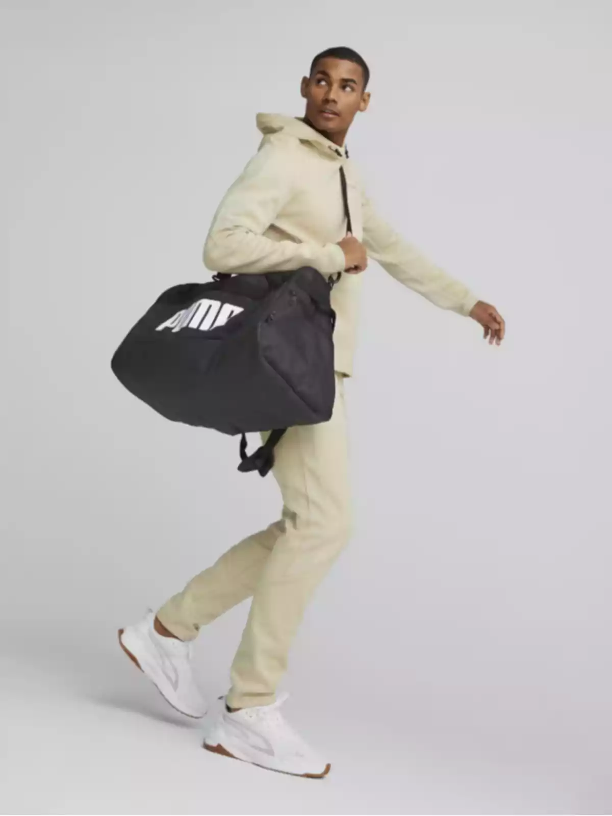 Hombre con sudadera y pantalón beige lleva una bolsa deportiva negra y mira hacia un lado mientras camina Hombre con sudadera y pantalón beige lleva una bolsa deportiva negra y mira hacia un lado mientras camina