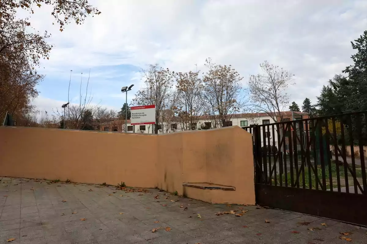 Entrada de una residencia con un muro color beige, una reja metálica y árboles sin hojas al fondo en un día nublado Entrada de una residencia con un muro color beige, una reja metálica y árboles sin hojas al fondo en un día nublado