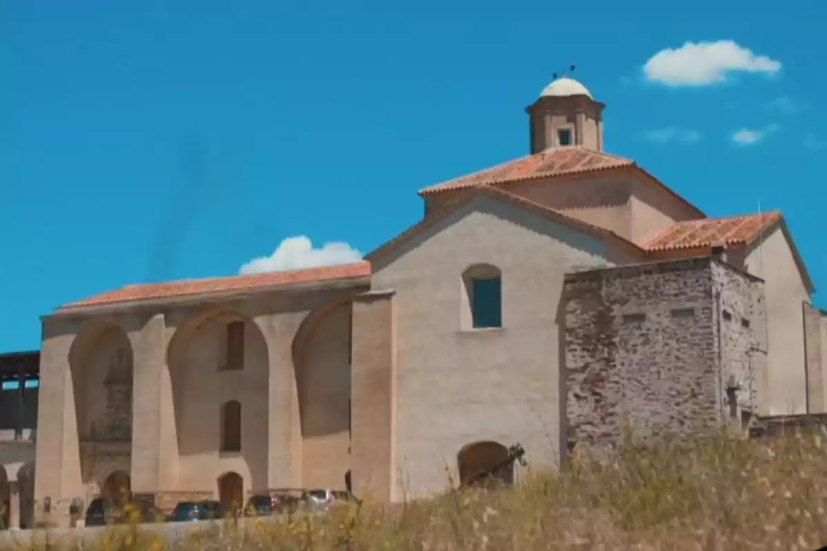 Edificio antiguo de paredes claras y techo de tejas rojas bajo un cielo azul con algunas nubes y vegetación en primer plano Edificio antiguo de paredes claras y techo de tejas rojas bajo un cielo azul con algunas nubes y vegetación en primer plano