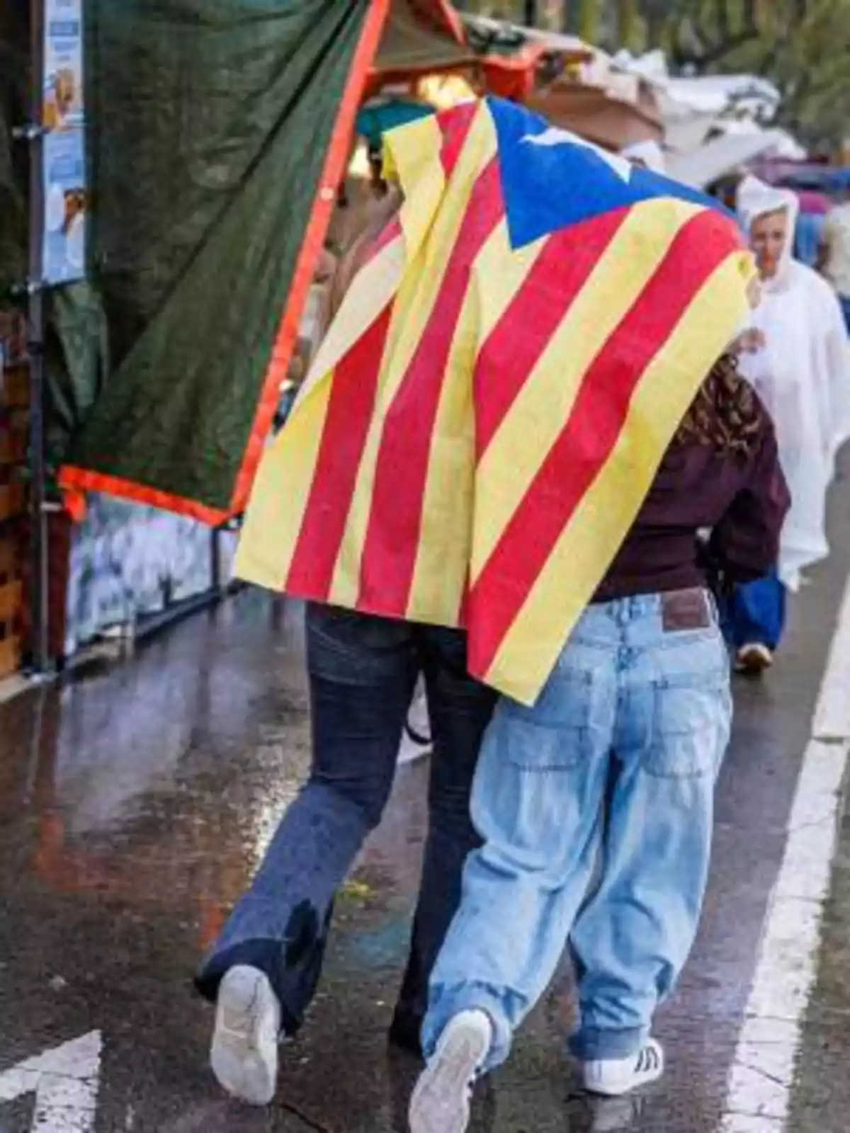 Dos personas caminan bajo la lluvia cubriéndose con una bandera estelada en una calle mojada