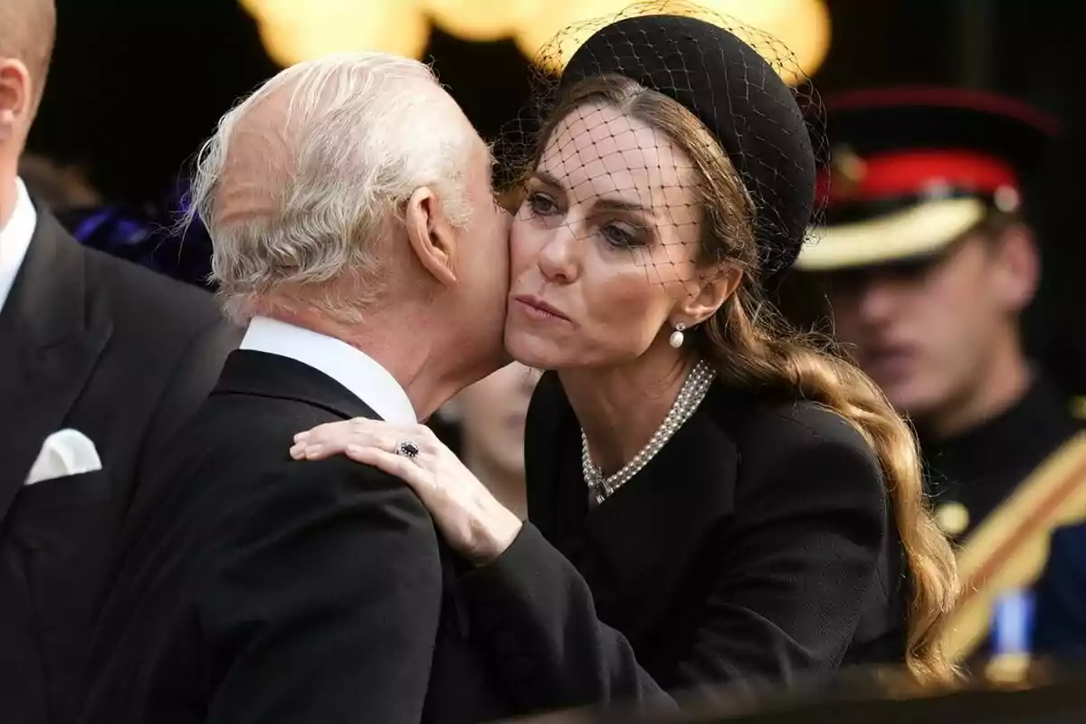 Una mujer vestida de negro con un tocado y velo abraza y besa en la mejilla a un hombre mayor de cabello canoso durante una ceremonia solemne Una mujer vestida de negro con un tocado y velo abraza y besa en la mejilla a un hombre mayor de cabello canoso durante una ceremonia solemne
