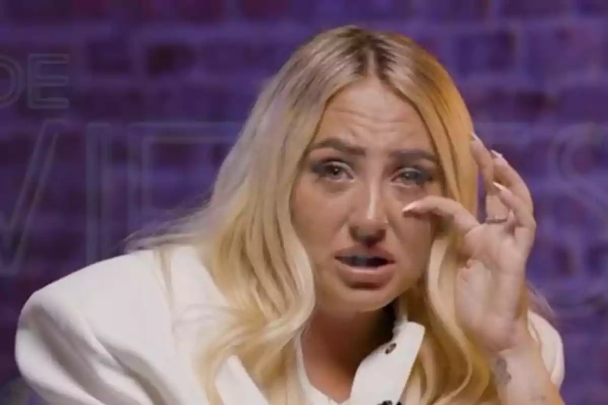 Rocío Flores rubia con expresión de tristeza y lágrimas en los ojos, vestida con saco blanco y fondo morado desenfocado Rocío Flores rubia con expresión de tristeza y lágrimas en los ojos, vestida con saco blanco y fondo morado desenfocado