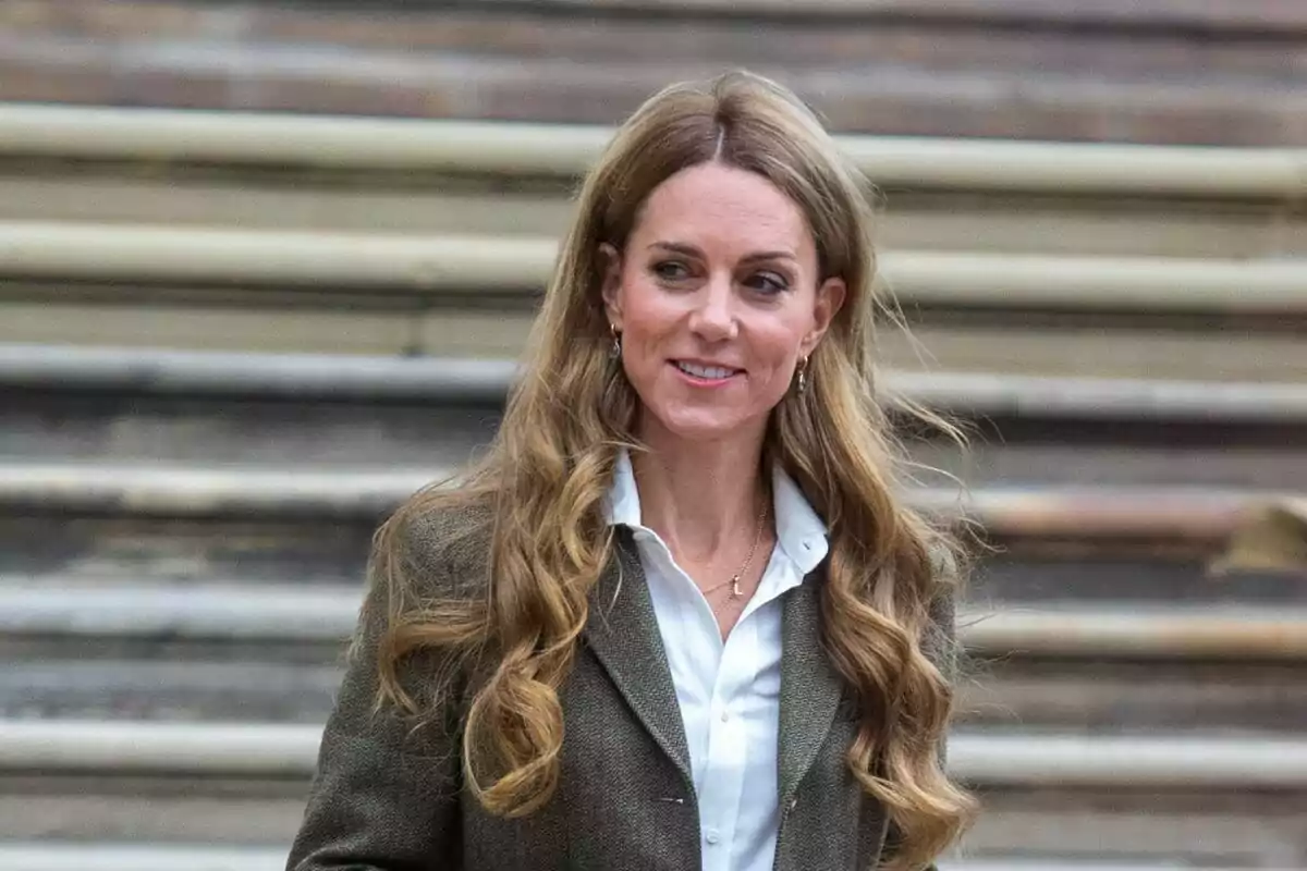 Mujer de cabello largo y ondulado con chaqueta marrón y camisa blanca de pie frente a unas escaleras