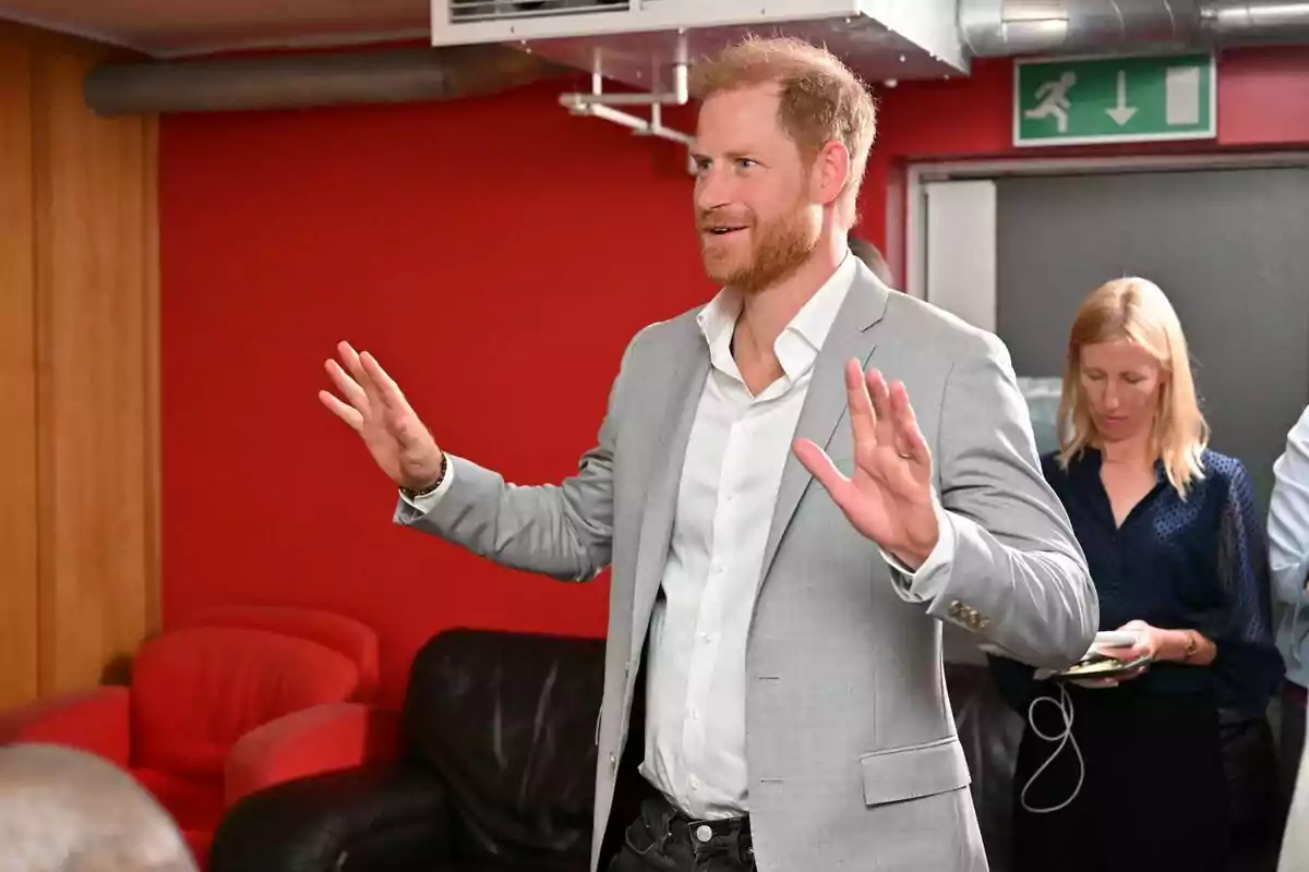 Hombre de cabello rubio y barba con saco gris y camisa blanca levantando las manos en una habitación con paredes rojas y muebles oscuros, mientras una mujer rubia está al fondo revisando algo