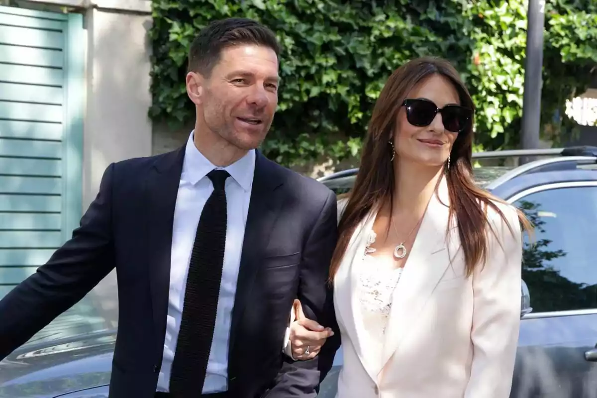 Pareja elegantemente vestida caminando al aire libre junto a un coche y vegetación de fondo