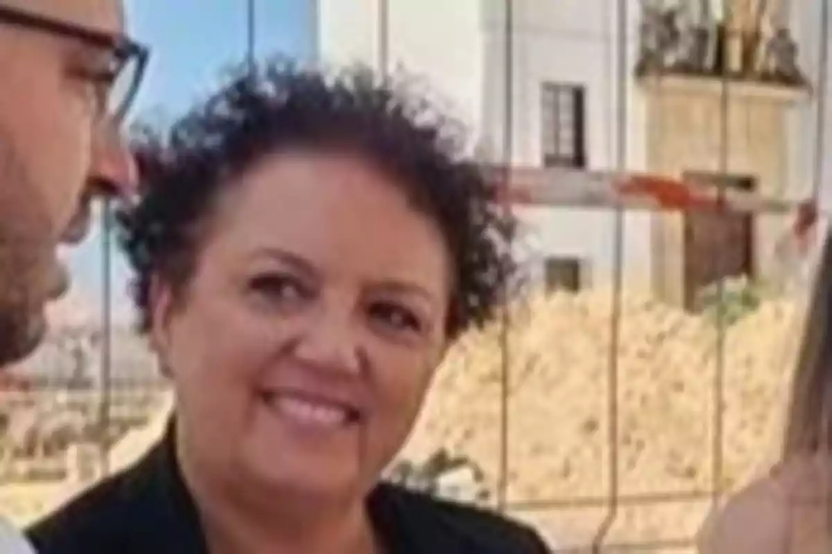 Mujer de cabello corto y rizado sonriendo mientras conversa con otras personas al aire libre frente a una construcción Mujer de cabello corto y rizado sonriendo mientras conversa con otras personas al aire libre frente a una construcción