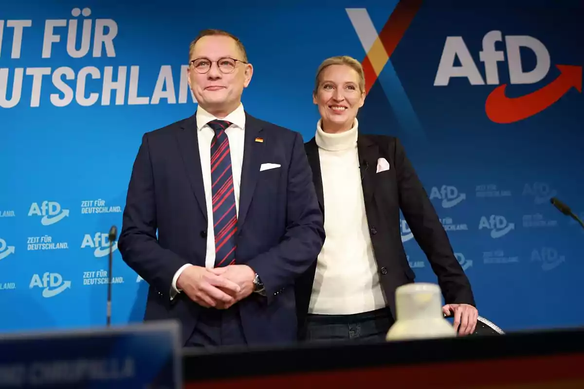 Dos personas vestidas de manera formal posan sonrientes frente a un fondo azul con logotipos y texto de AfD Dos personas vestidas de manera formal posan sonrientes frente a un fondo azul con logotipos y texto de AfD