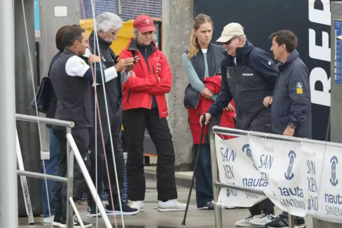 Un grupo de personas vestidas con ropa deportiva y abrigos conversan y observan algo en un puerto junto a una barandilla y una lona con logotipos de un evento náutico