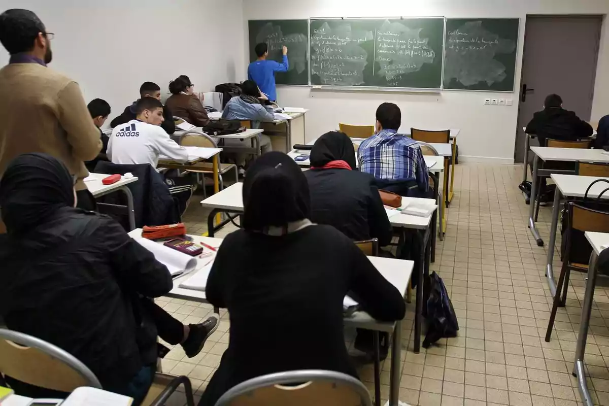 Estudiantes sentados en pupitres prestan atención mientras una persona escribe en el pizarrón y otra observa de pie al fondo del aula
