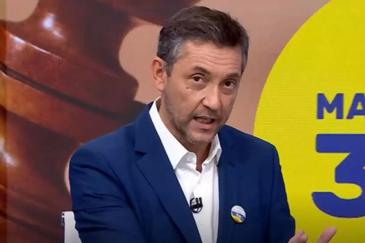 Hombre de cabello corto y barba con saco azul y camisa blanca hablando en un set de televisión con fondo colorido y un pin en la solapa Hombre de cabello corto y barba con saco azul y camisa blanca hablando en un set de televisión con fondo colorido y un pin en la solapa