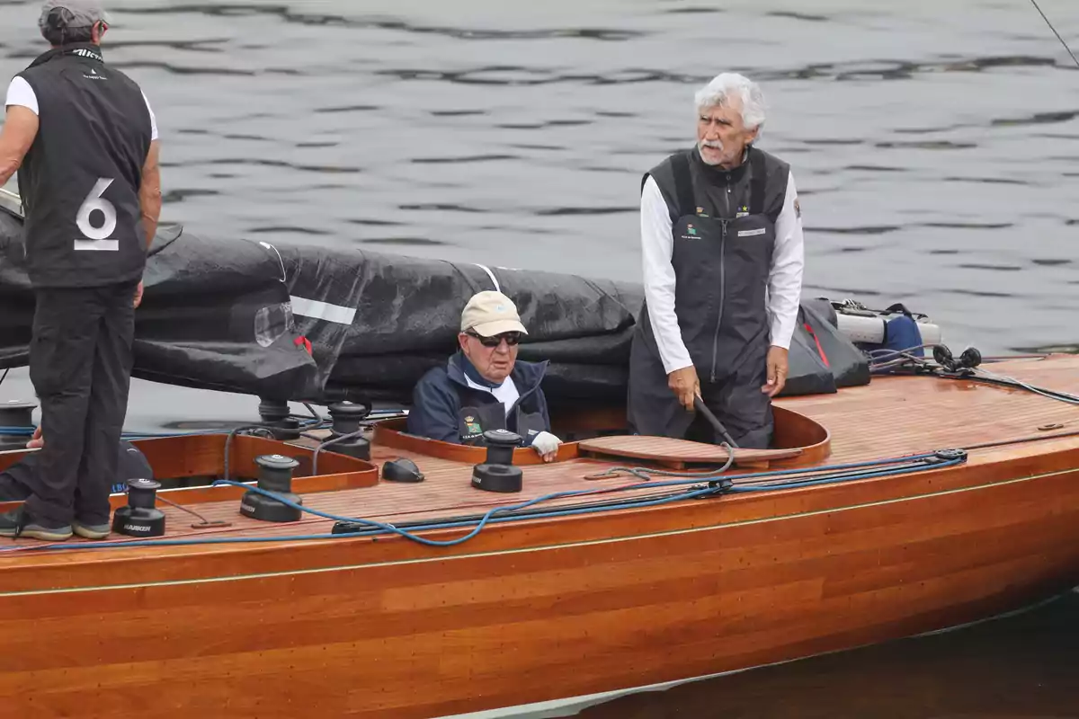 Juan Carlos I y dos hombres más navegando en un velero de madera sobre aguas tranquilas, uno de ellos lleva gorra y gafas de sol, otro sostiene el timón y el tercero está de pie con el número 6 en la espalda