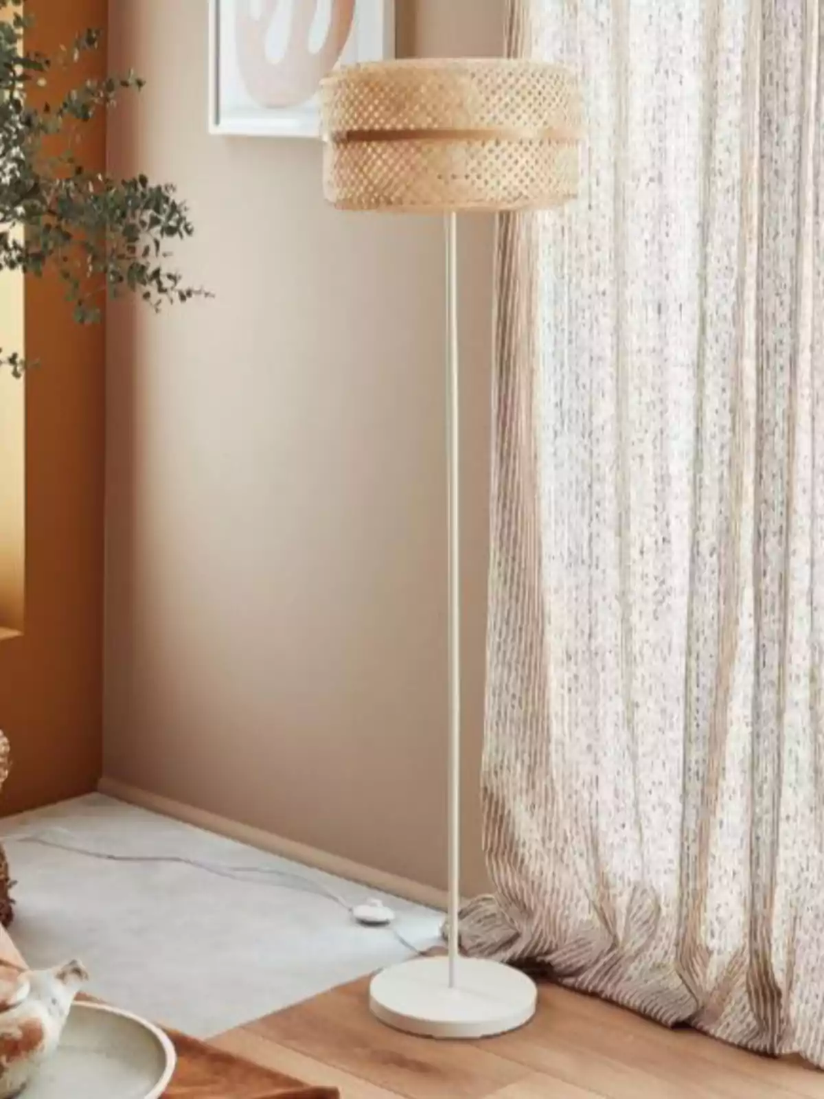 Lámpara de pie con base y estructura blanca y pantalla de mimbre en una sala decorada en tonos neutros junto a una cortina clara