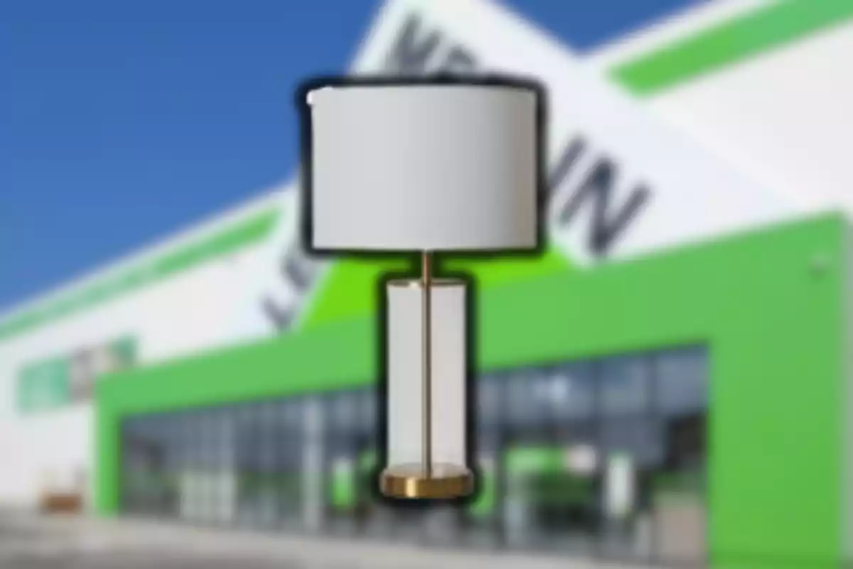 Lámpara de mesa moderna con base dorada y pantalla blanca frente a una tienda con fachada verde y blanca desenfocada