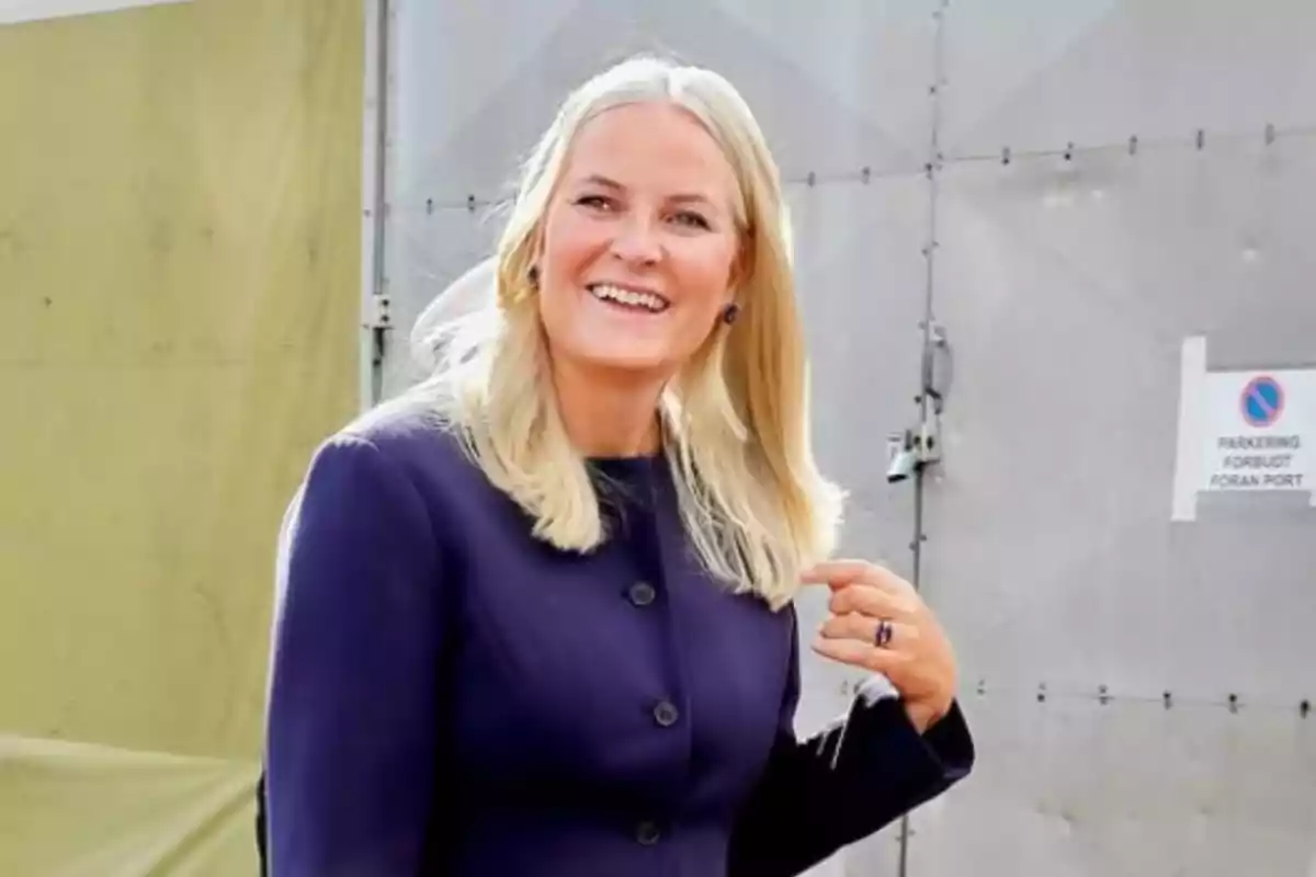 Mette-Marit, sonriente con chaqueta morada de pie al aire libre frente a una pared metálica