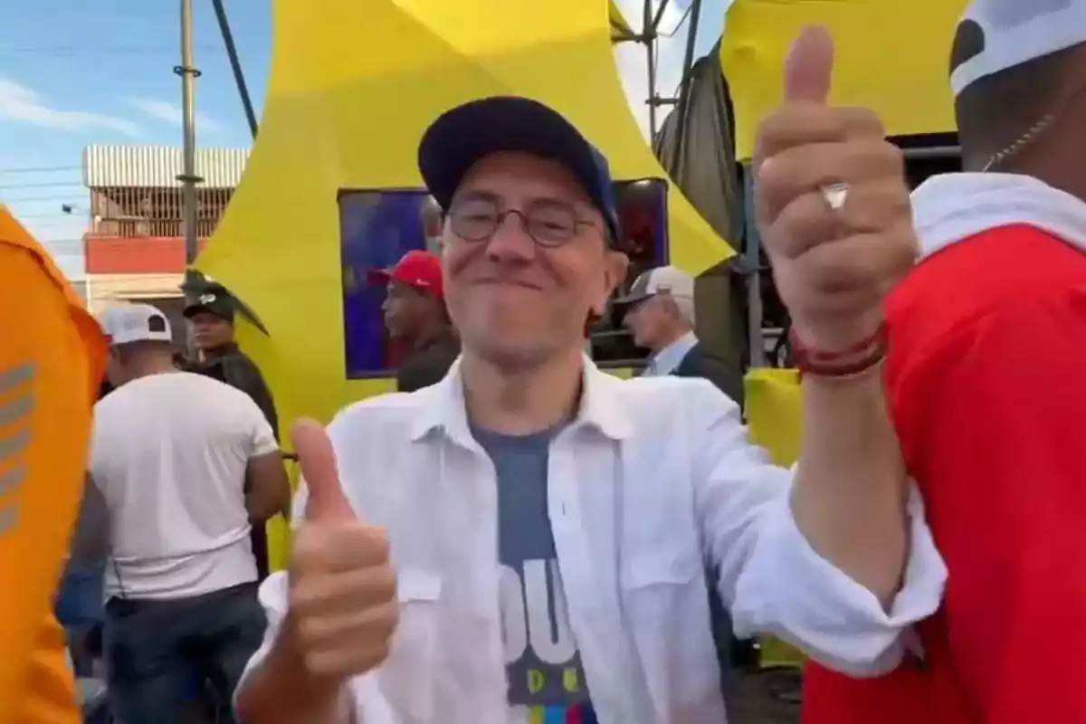 Hombre con gafas y gorra azul sonríe y muestra ambos pulgares hacia arriba en un evento al aire libre con fondo amarillo y varias personas alrededor Hombre con gafas y gorra azul sonríe y muestra ambos pulgares hacia arriba en un evento al aire libre con fondo amarillo y varias personas alrededor