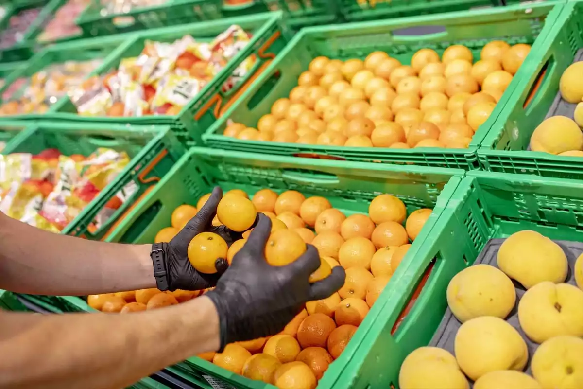 Persona con guantes negros seleccionando naranjas en un supermercado con cajas verdes llenas de frutas Persona con guantes negros seleccionando naranjas en un supermercado con cajas verdes llenas de frutas