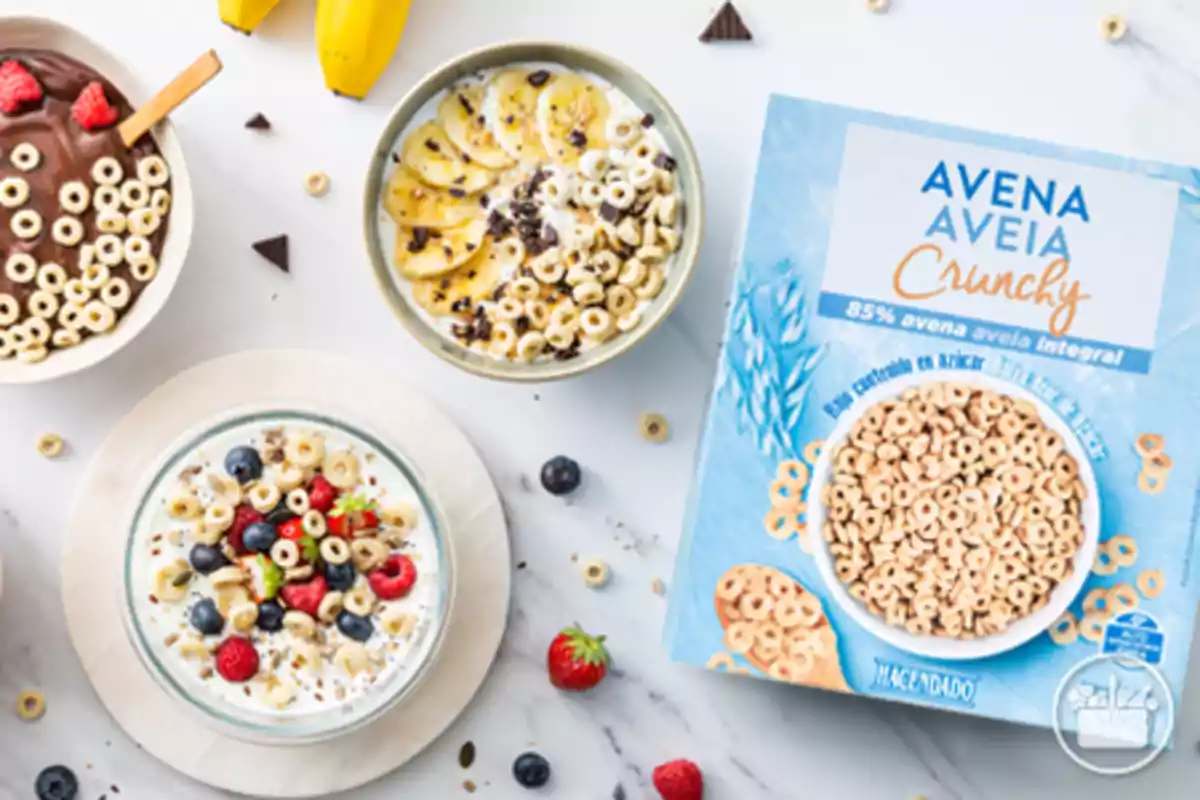 Cereal de avena en forma de aros servido en diferentes bowls con frutas y yogur junto a una caja azul de Avena Crunchy de la marca Hacendado sobre una mesa blanca