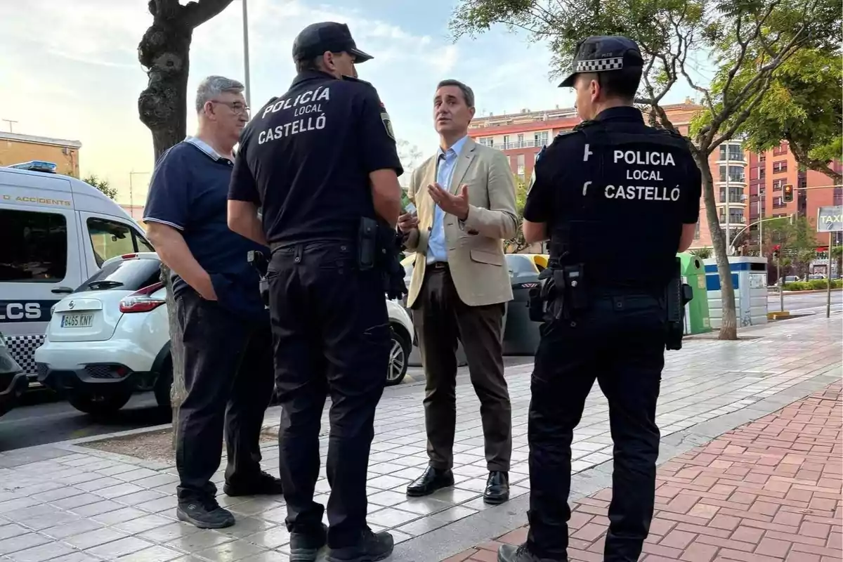 Cuatro personas conversan en una acera, dos de ellas son policías locales de Castelló y al fondo se ven vehículos y edificios urbanos.