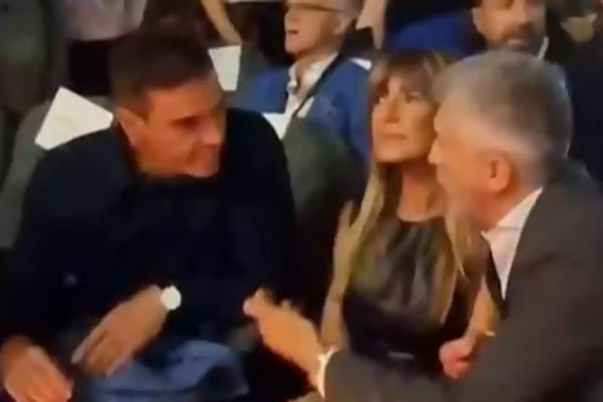 Tres personas sentadas conversando en un evento social con más gente al fondo Tres personas sentadas conversando en un evento social con más gente al fondo