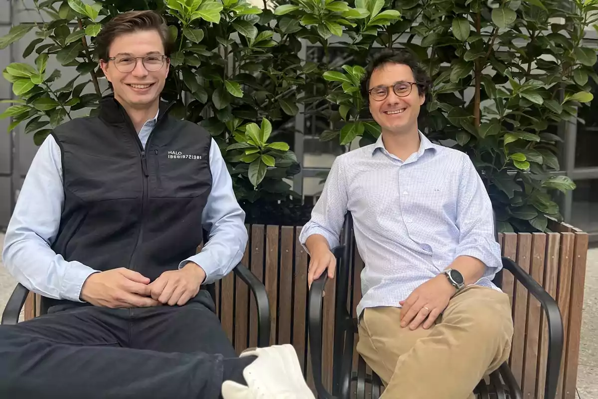 Dos hombres jóvenes sentados en una banca de madera al aire libre sonriendo frente a unas plantas verdes