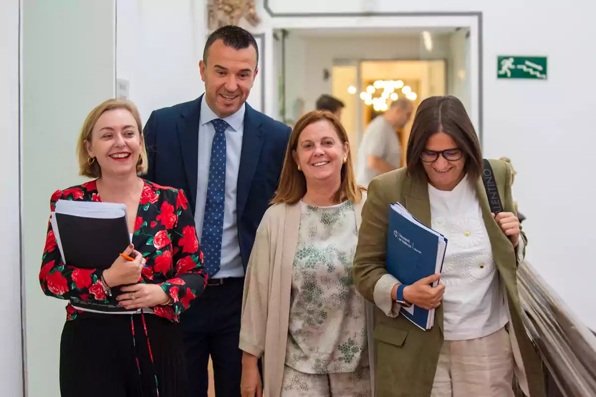 Cuatro personas sonríen mientras caminan por un pasillo, dos de ellas llevan carpetas y documentos en las manos Cuatro personas sonríen mientras caminan por un pasillo, dos de ellas llevan carpetas y documentos en las manos