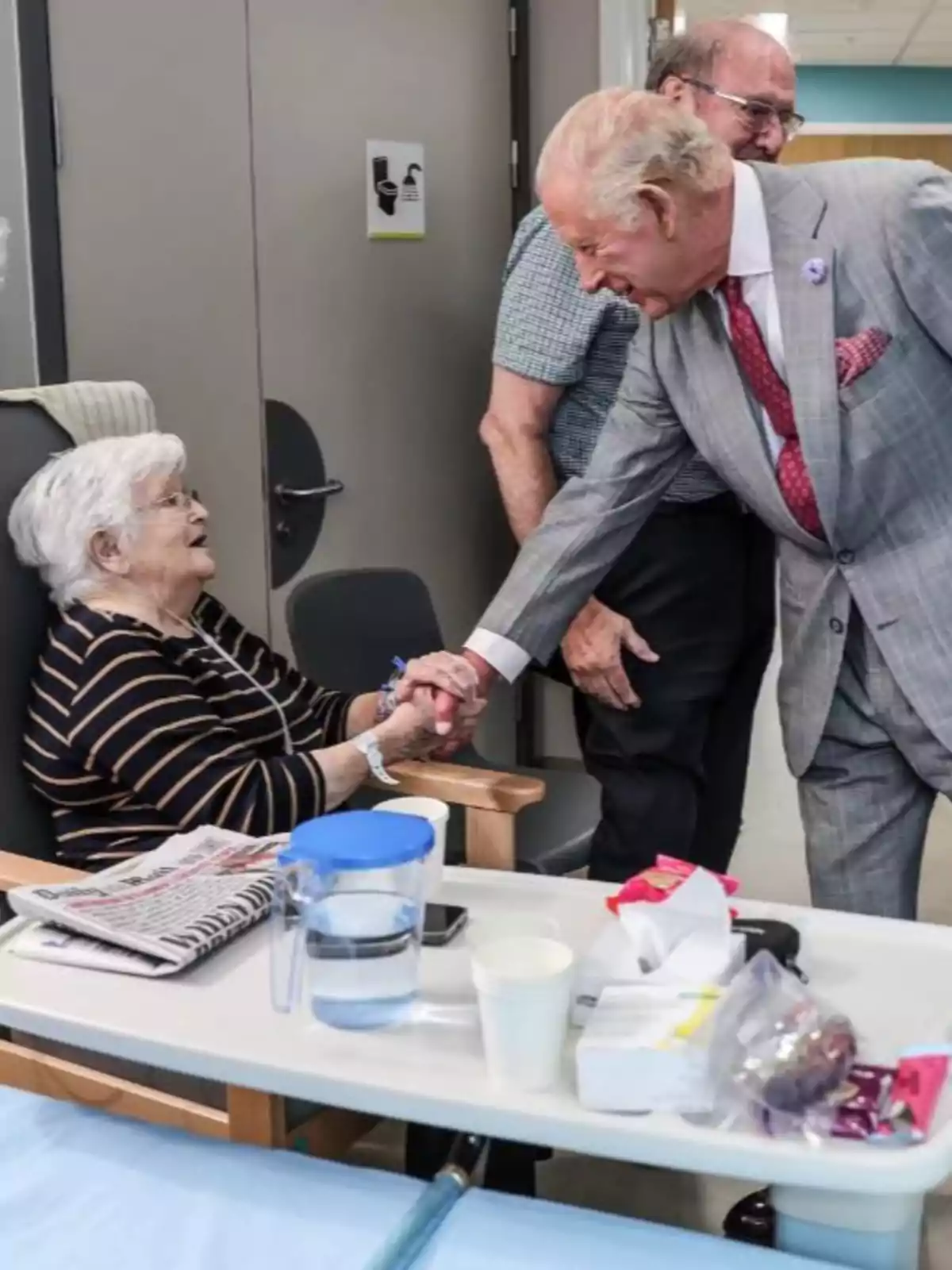 King Charles III, elegantly dressed, greets an elderly woman sitting in a chair inside a room, while another person watches them and a table with objects is in the foreground. King Charles III, elegantly dressed, greets an elderly woman sitting in a chair inside a room, while another person watches them and a table with objects is in the foreground.